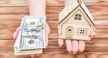 Woman's hands holding money and model of house over wooden table.