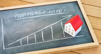 Rising Interest Rates Graph representing the rise in mortgage interest rates drawn on a chalkboard lying on a wooden table. A model of a house with a red roof is on the chalkboard. Finance and real estate concept.
