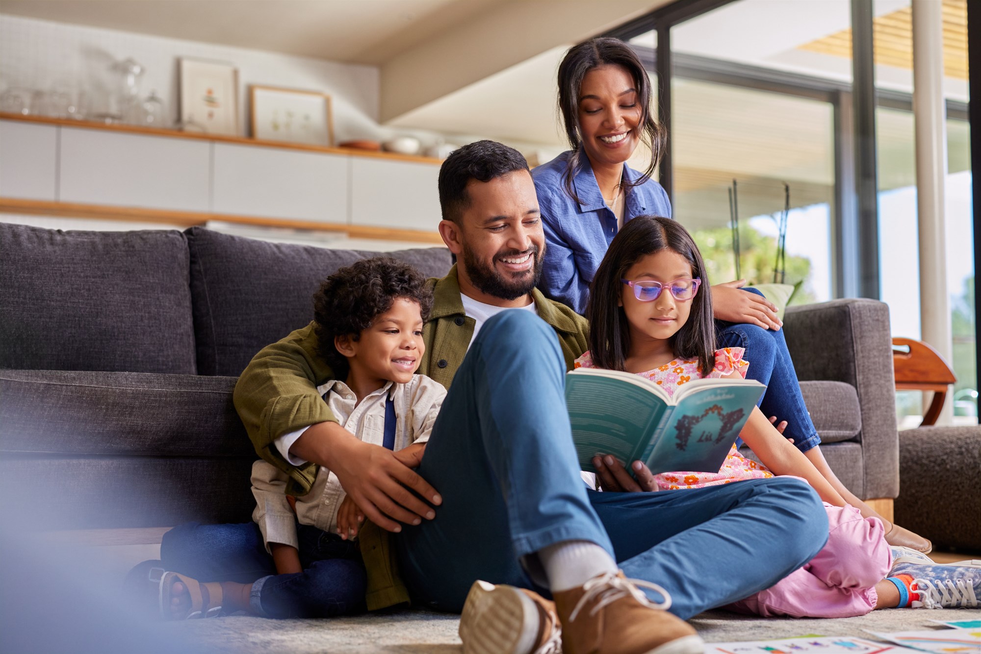 Mixed race father reading a story book to daughter and son while sitting with beautiful wife on couch. Young multiethnic man sitting on floor carpet reading a fairy tale to his kids. Happy parents holding book and spending time with children on a happy weekend.