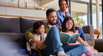 Families, Kids & Quality of Life Mixed race father reading a story book to daughter and son while sitting with beautiful wife on couch. Young multiethnic man sitting on floor carpet reading a fairy tale to his kids. Happy parents holding book and spending time with children on a happy weekend.