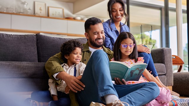 Mixed race father reading a story book to daughter and son while sitting with beautiful wife on couch. Young multiethnic man sitting on floor carpet reading a fairy tale to his kids. Happy parents holding book and spending time with children on a happy weekend.